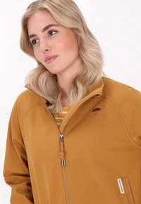 Rudas zip-up striukė su aukšta apykakle ir siuvinėtu skėčio logotipu. Apima pilkus užtrauktukus ir vidinę dryžuotą marškinėlių. 