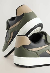 Basket en vert et noir avec des accents beiges, en matériau synthétique lisse, à bout rond, avec une sangle Velcro, et une semelle en caoutchouc blanc avec un profil noir.