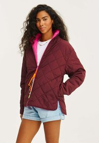 Burgundská prošívaná bunda s růžovou podšívkou, s oranžovým zipem a dekorativními táhly a bočními rozparky. Nositelská přes bílou košili.