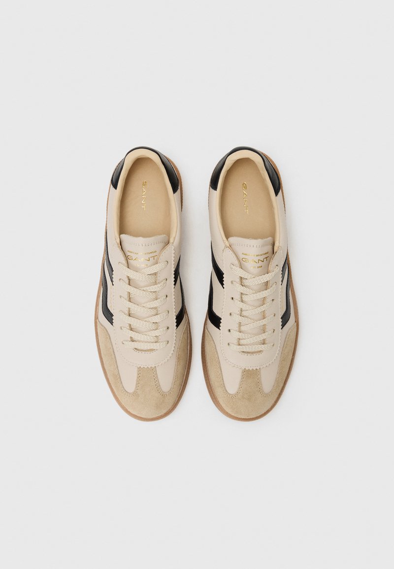 Beige Sneaker mit schwarzen Seitenstreifen, Wildlederakzenten und Schnürverschluss. Verfügt über eine braune Gummisohle und ein geprägtes Logo auf der Zunge.