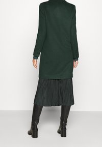 Cappotto lungo di lana verde scuro con colletto e polsini abbottonati, abbinato a una gonna nera plissettata e stivali neri fino al ginocchio.