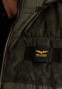 Veste en velours côtelé vert foncé avec boucle noire affichant le logo jaune "PME Legend Tested at High Altitude" et un emblème d'aile.