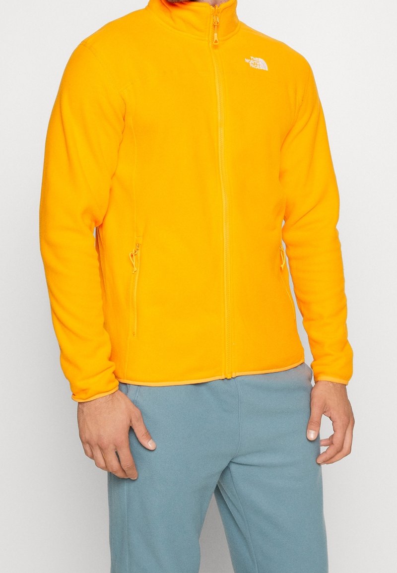 Hombre vestido con una chaqueta de forro polar naranja brillante con cremalleras en los bolsillos laterales y pantalones azules claros, de pie con los brazos relajados.