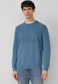 Pull bleu clair avec des motifs en maille texturée, un col rond et des poignets côtelés. Présente une petite étiquette de marque sur la poitrine.