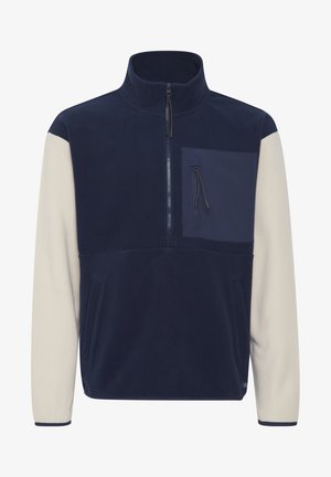 Pull en polaire bleu marine avec manches crème. Comprend un col zippé, des poches latérales et une poche sur la poitrine avec zip. Texture douce, design moderne.