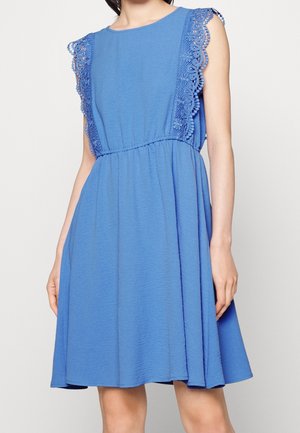 Robe bleue sans manches avec bordure en dentelle florale sur les épaules, taille cintrée et jupe évasée portée par une femme sur un fond uni.