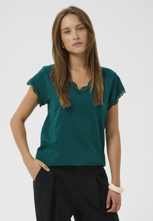 Mujer con el pelo largo y castaño, vistiendo una camiseta de manga corta color azul verdoso con detalles de encaje y pantalones negros, de pie con una mano en el bolsillo y expresión neutral.