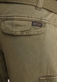 Olive-zielone spodnie cargo z tkaniny, posiadające tylną kieszeń z przeszywaną klapą, teksturowany materiał oraz małą metkę marki w pobliżu kieszeni.