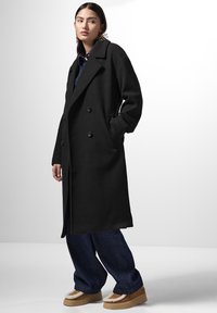 Cappotto in lana nera doppiopetto con ampi revers e tasche laterali, abbinato a pantaloni in denim a gamba larga e scarpe con plateau beige.