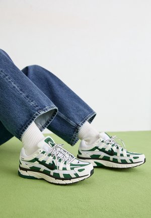Personne portant des chaussures de course Nike blanches et vertes avec des chaussettes blanches et un jean bleu, assise sur un sol vert contre un mur blanc.