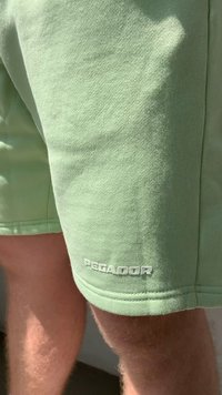 Žalios medvilnės šortai, pasižymintys lygia tekstūra ir balta "PEGADOR" logo netoli apačios, su laisvu střilimu ir vidutinio ilgio iki šlaunų.