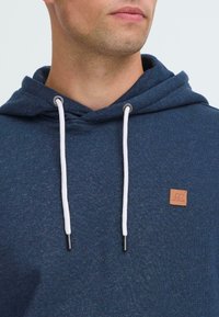 Uomo che indossa un felpa blu con lacci bianchi e un piccolo patch in pelle marrone sul petto, che guarda leggermente verso destra.