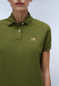 Zelené polo tričko s texturovanou tkaninou, dvě bílé knoflíky na límci a malým zlatým logem s designem vlajky na hrudi.