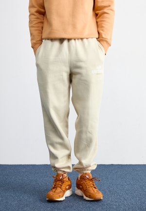 Person trägt beige Sweatpants, orange Sneaker und ein hellbraunes Sweatshirt mit den Händen in den Taschen, steht auf blauem Teppich.