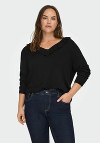 Žena s dugom smešnom kosom nosi crni pulover s nabranim V-izrezom i tamnoplave traperice, pozira s jednom rukom u džepu.