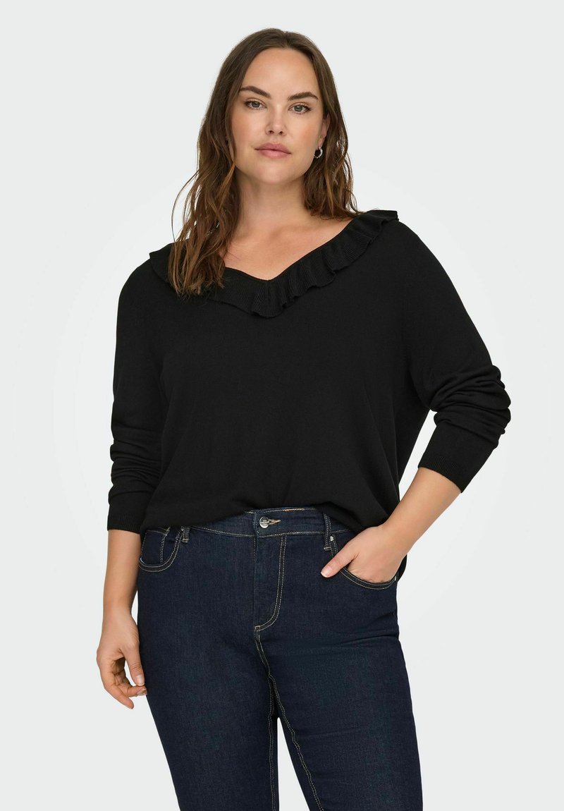 Žena s dugom smešnom kosom nosi crni pulover s nabranim V-izrezom i tamnoplave traperice, pozira s jednom rukom u džepu.