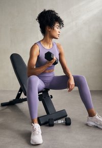 Vijolična športna obleka, vključno s tesnim topom in leggins hlačami, v kombinaciji s svetlimi supergami. Drži črno utež. V bližini je nastavljiva utežna klop.
