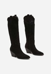 Bottes noires en daim hauteur genou avec bouts pointus, talons compensés, découpes décoratives et détails de couture. Fermeture éclair sur le côté.