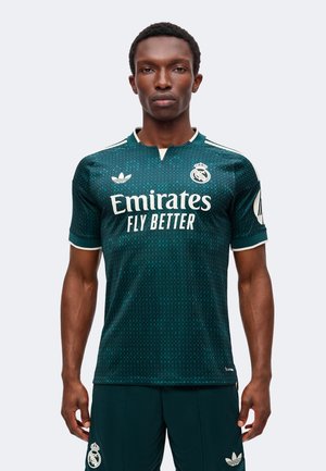 Männliches Model trägt dunkelgrünes Real Madrid Fußballtrikot und passende Shorts mit Emirates Fly Better Sponsorlogo, steht vor neutralem Hintergrund.