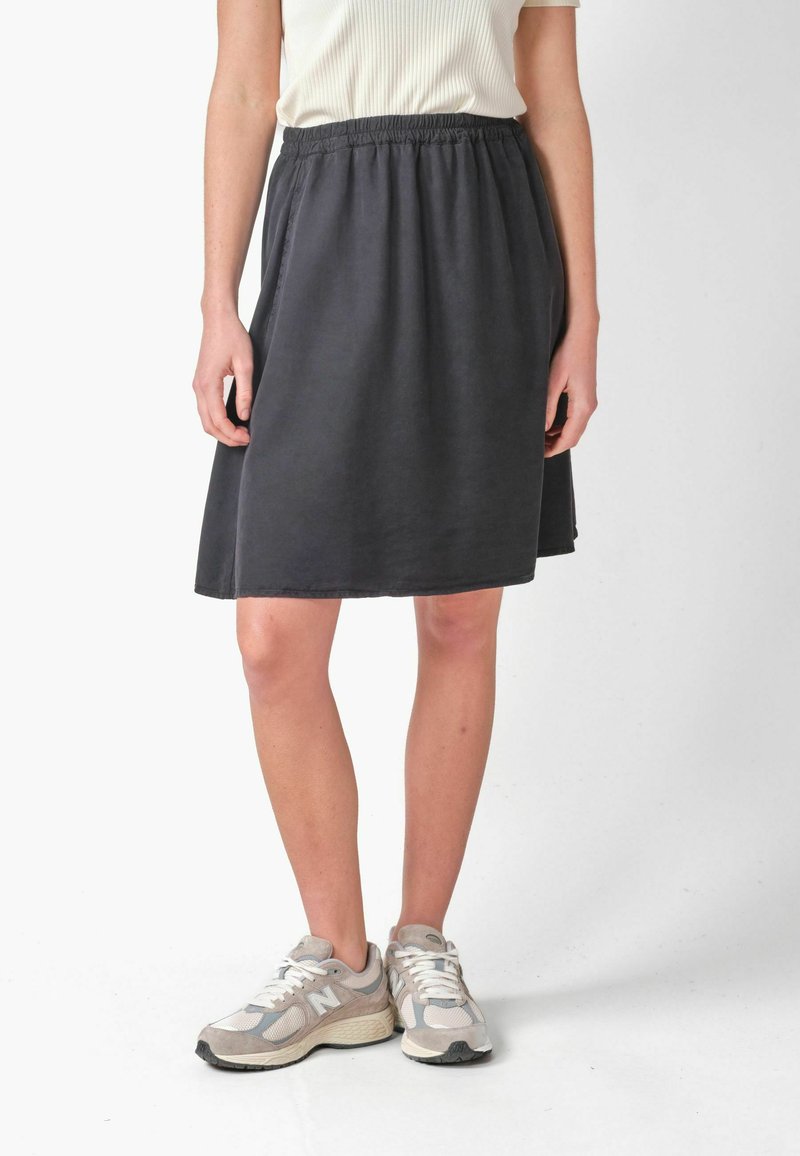 Personne portant une jupe grise foncée longueur genou avec taille élastique et baskets blanches, debout devant un fond clair uni.