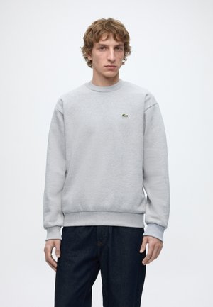 Ung mand med bølget hår iført en lysegrå crewneck sweatshirt og mørke jeans foran en ensfarvet hvid baggrund.