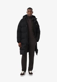 Abrigo acolchado negro con diseño largo, cierre de cremallera y capucha, combinado con un suéter oscuro, pantalones marrones y zapatillas.