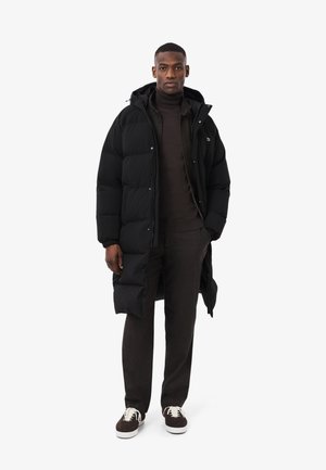 Schwarzer Steppmantel mit langem Schnitt, Reißverschlussverschluss und Kapuze, kombiniert mit einem dunklen Pullover, brauner Hose und Sneakers.