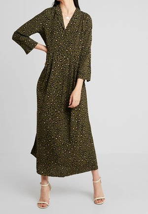 Kobieta ubrana w oliwkową, długorękawową sukienkę midi w panterkę, stojąca z jedną ręką na biodrze i beżowymi szpilkami z paskiem wokół kostki.