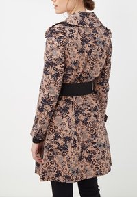 Trench de flores en rosa claro con diseños en negro y azul marino. Presenta un cuello ancho, mangas largas y una cintura ceñida con cinturón para mayor definición.