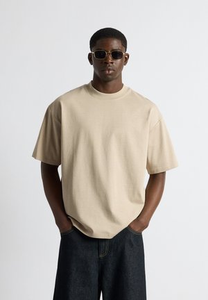 Mand iført beige oversized t-shirt, mørke jeans og mørke solbriller, stående med hænderne i lommerne mod en ensfarvet lys baggrund.