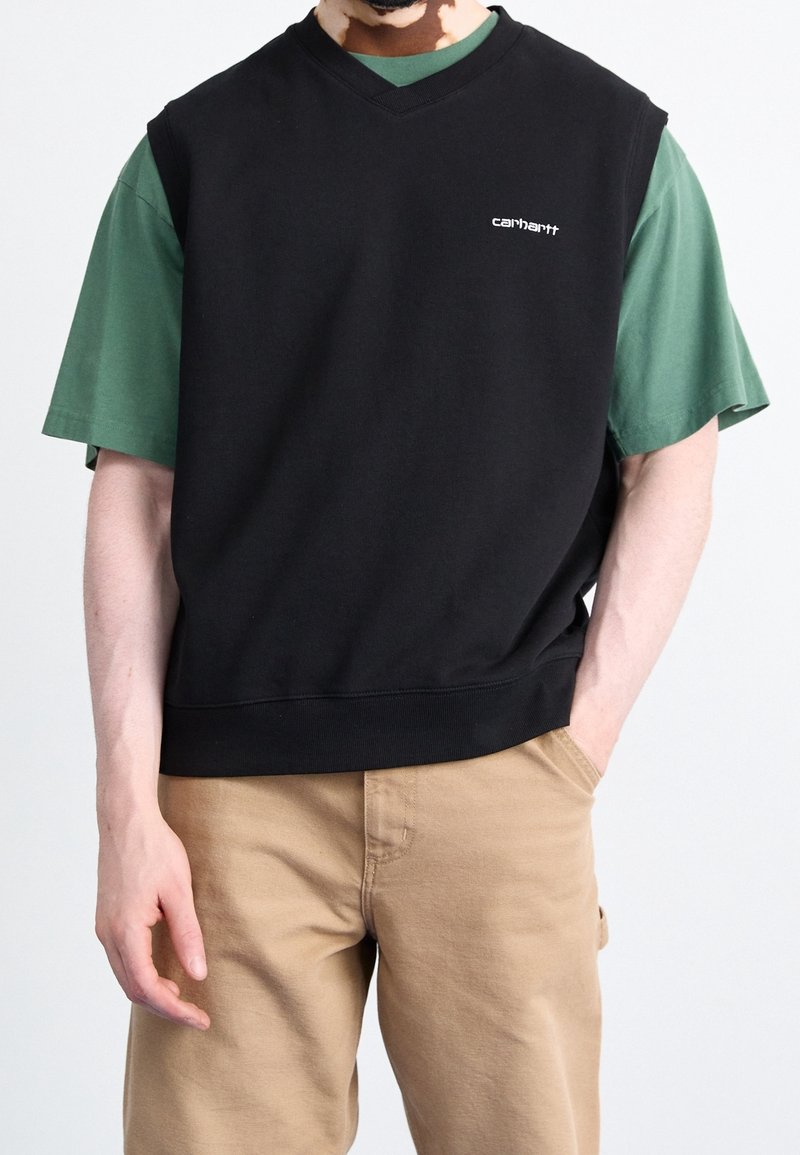 Felpa nera senza maniche con scollo a V, indossata sopra una t-shirt verde. Presenta un logo bianco sul petto e un orlo a coste. Pantaloni beige visibili.