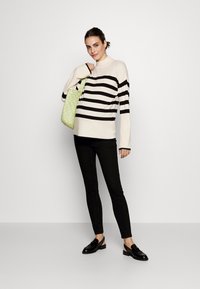 Maglione crema con righe nere, pantaloni slim-fit neri e mocassini neri. Tiene una borsa a tracolla intrecciata verde. Design semplice e look moderno.
