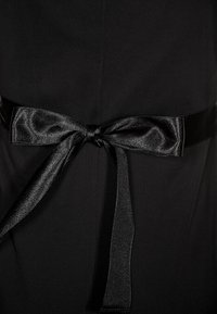 Mustan mekko, jossa on sileä kangas ja leveä satiinivyö, joka on solmittu kauniiksi nauhaksi vyötärölle. Vyössä on hienovarainen kiilto ja strukturoitu pinta.