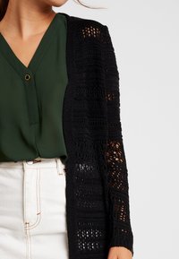 Black knitted cardigan with openwork pattern, featuring long sleeves, textured fabric, and a loose fit, paired with a green blouse.
