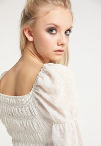 White textured blouse with puff sleeves, featuring smocked detailing and a square neckline. Soft fabric with small raised dots.