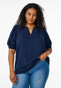 Navy blue pusero, jossa on puffihihat, teksturoitu pisaramainen kuvio, V-pääntie ja korostusompeleet, yhdistettynä tiukkoihin sinisiin farkkuihin.