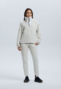 Nainen seisoo päällään vaalea beige fleece-takki, jossa on vetskari, beige housut ja mustat lenkkarit, neutraalia vaaleaa taustaa vasten.
