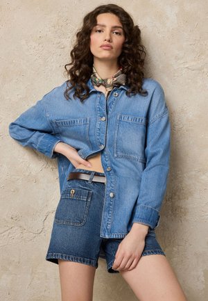 Ung kvinde med krøllet hår har en blå denimjakke, mørke denimshorts med bælte og et mønstret tørklæde omkring halsen.