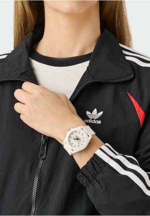 Balta rankinė laikrodžio apyrankė su aštuonkampiu ciferblatu, juodais skaičiais ir spalvingomis rodyklėmis, dėvima ant rankos, kartu su juodu švarku su baltomis juostelėmis.