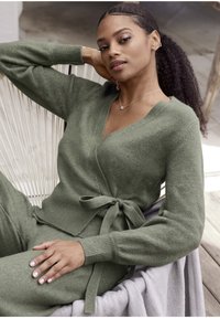 Cardigan verde avvolgente, materiale in maglia morbido, scollo a V, maniche lunghe, dettaglio con cintura, indossato sopra pantaloni coordinati, con uno sfondo testurizzato.