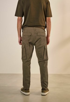 Pantalones cargo en algodón verde oliva, con múltiples bolsillos laterales y un diseño de pierna ajustada. Combinados con una camiseta oscura y zapatillas.