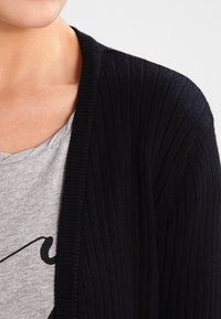 Schwarzer gerippter Cardigan mit offenem Design, aus Strickmaterial gefertigt, über einem grauen T-Shirt mit einem schlichten Grafikmotiv getragen.