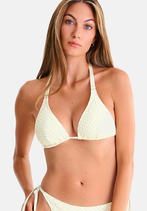 Jeune femme aux longs cheveux blonds portant un haut de bikini halter texturé blanc cassé et un bas de bikini assorti avec des liens sur les côtés sur un fond uni.