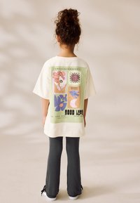 T-shirt oversize couleur crème avec un motif graphique multicolore au dos, associé à un pantalon gris évasé. Matière en coton.
