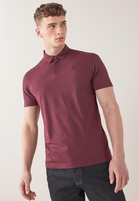 Paita polo maroon dengan lengan pendek terbuat dari katun, dilengkapi dengan placket tiga kancing dan logo hitam kecil di dada.
