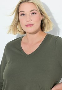 Olivgrön V-ringad topp i slät tyg med långa ärmar, med en avslappnad passform och subtil textur. Guldörhängen i form av hoop som en framträdande accessoar.