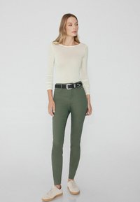 Un haut blanc transparent à manches longues associé à un pantalon olive bien coupé et une ceinture noire. Les chaussures sont des baskets claires avec une semelle beige.