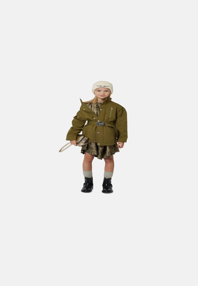 Olijfgroen oversized jack met een riem, gelaagd over een gouden jurk, gecombineerd met grijze kniekousen en zwarte enkellaarsjes; crèmekleurige gebreide beanie.