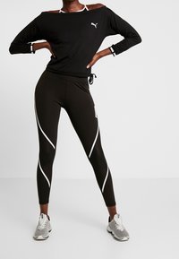 Black off-shoulder long-sleeve top with white logo, paired with black leggings featuring white curved stripes and grey athletic shoes.