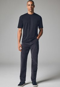 Sort bomulds t-shirt med korte ærmer, parret med mørkegrå kordfløjl bukser. Mand der står afslappet med hænderne i lommerne og iført mørke sneakers.
